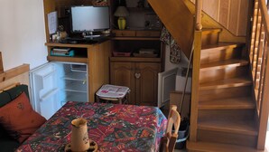 A compact room with a bed, a desk, a small table, a refrigerator, a wooden staircase, and a clock on the wall.