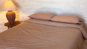 Una cama con una manta y almohadas color beige, una mesita de noche redonda de madera con una lámpara, y una pared blanca con textura.