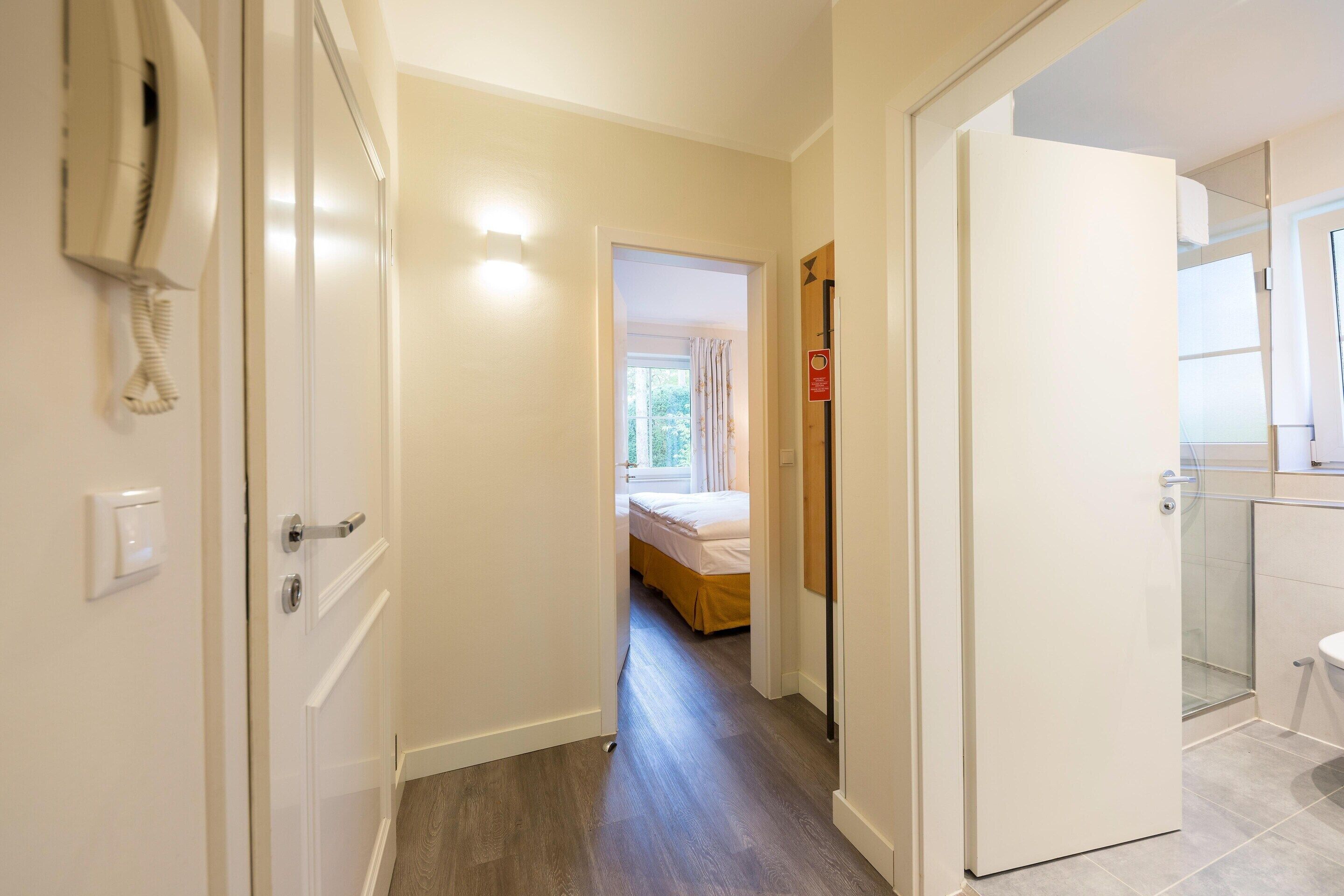 A narrow hallway with a bed at the end, a bathroom with a toilet and shower, and a phone on the wall.