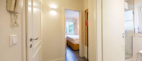 A narrow hallway with a bed at the end, a bathroom with a toilet and shower, and a phone on the wall.