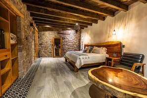 A cozy bedroom with a stone wall, wooden beams, a bed with pillows, a chair, and a wooden coffee table. - Hotel Palacio Del Gambusino (Real de Catorce)