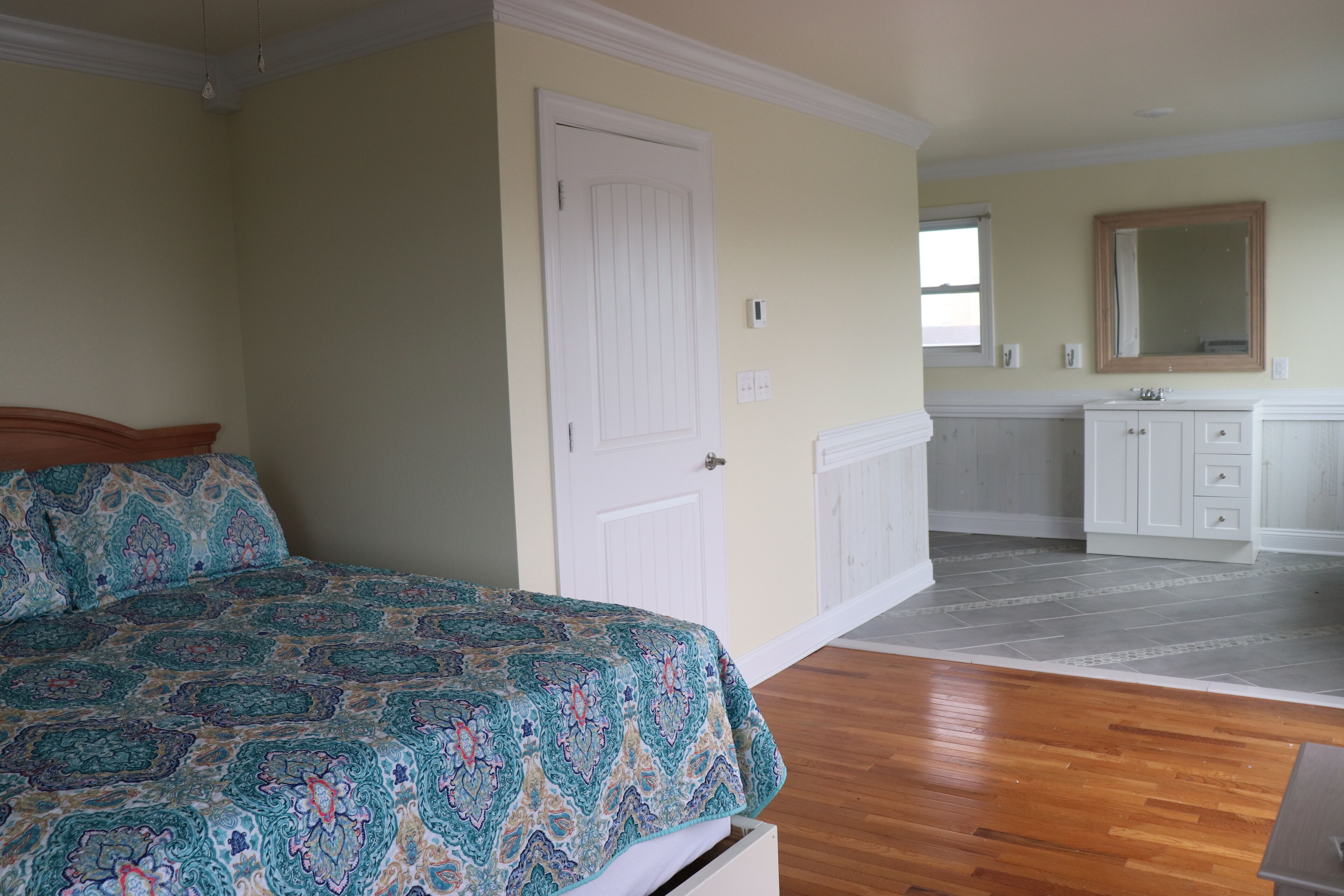 A bedroom with a bed, a white door, a bathroom with a sink and mirror, and a hallway with a wooden floor.