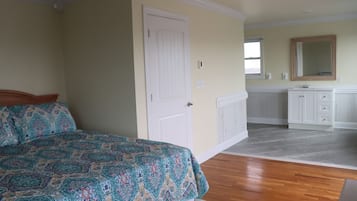 A bedroom with a bed, a white door, a bathroom with a sink and mirror, and a hallway with a wooden floor.