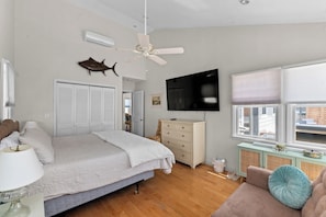 A bedroom with a bed, a ceiling fan, a TV, a dresser, a window with blinds, and a view of the outside.