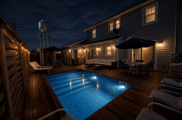A swimming pool with lounge chairs and a barbecue area, surrounded by a wooden deck and a house with exterior lighting.