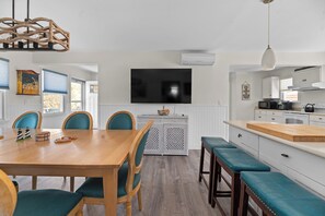 A modern kitchen with white cabinetry, a large flat-screen TV, and a dining area with wooden table and teal chairs.