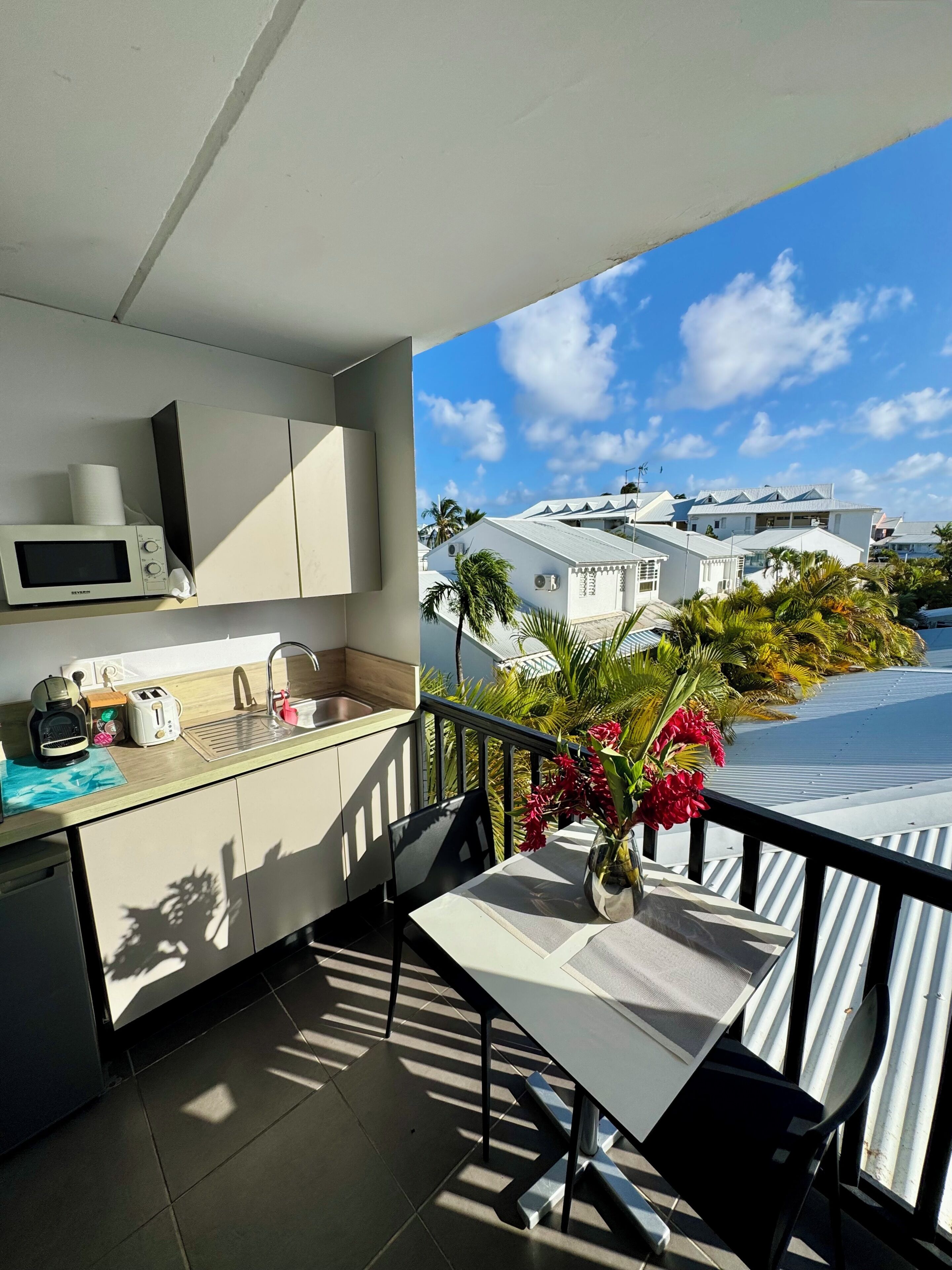 A balcony with a view of neighboring houses, a small kitchen with a microwave, toaster, and sink, and a table with a vase of flowers.