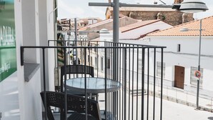 A balcony with a table and chairs, overlooking a street with buildings and a tiled roof.