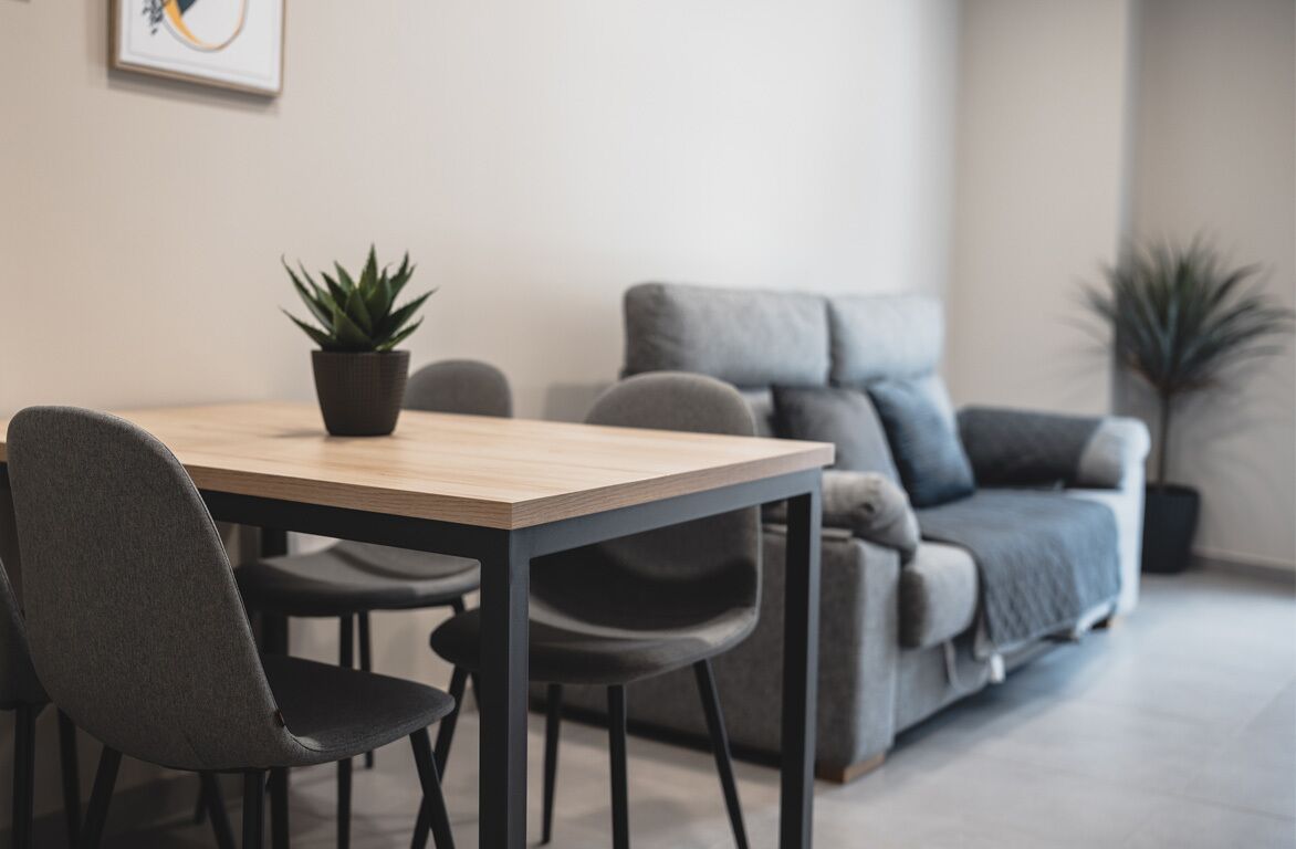 A modern living room with a wooden dining table, grey upholstered chairs, a grey sofa, and two potted plants.