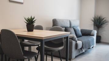 A modern living room with a wooden dining table, grey upholstered chairs, a grey sofa, and two potted plants.