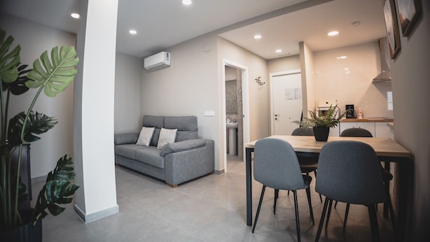 A modern living room with a grey sofa, a dining table with chairs, and a kitchen area in the background.
