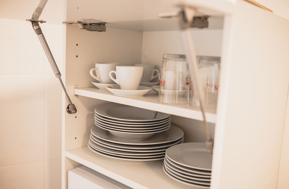 Un armario de cocina con estantes blancos que contienen tazas, platillos y platos apilados.