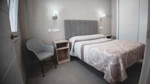A small, neatly arranged bedroom with a single bed, a chair, a nightstand, and two wall-mounted lights.