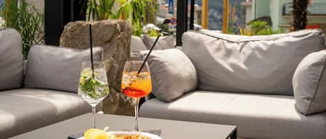 A modern outdoor seating area with a sofa and a table. There are two drinks on the table, one with a straw and one without. A plate with food and a napkin is also on the table.