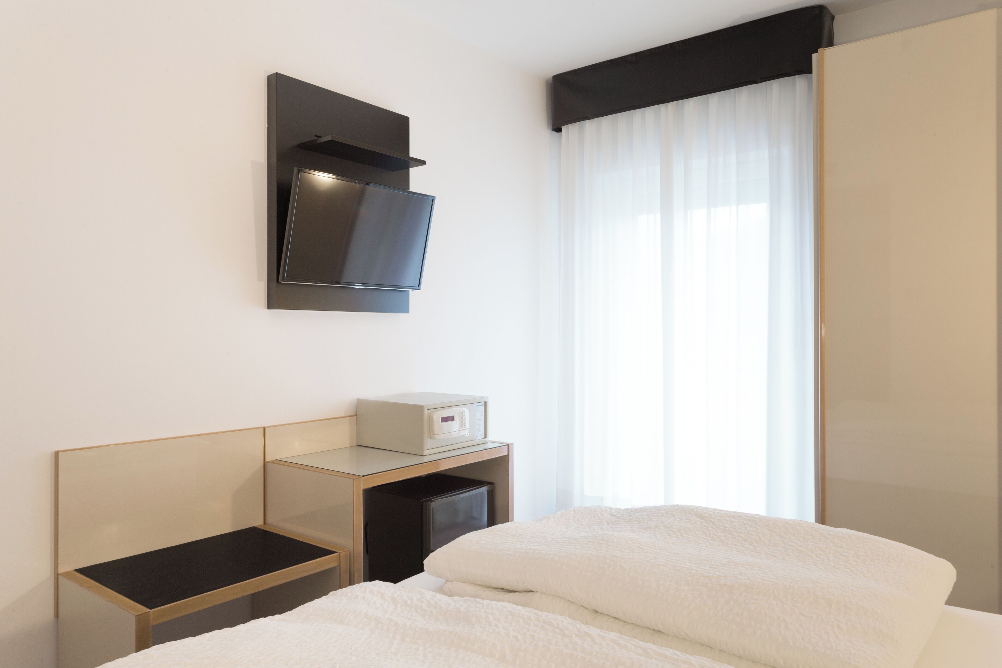 Una habitación de hotel con cama, mesita de noche, televisión montada en la pared y ventana con cortinas.
