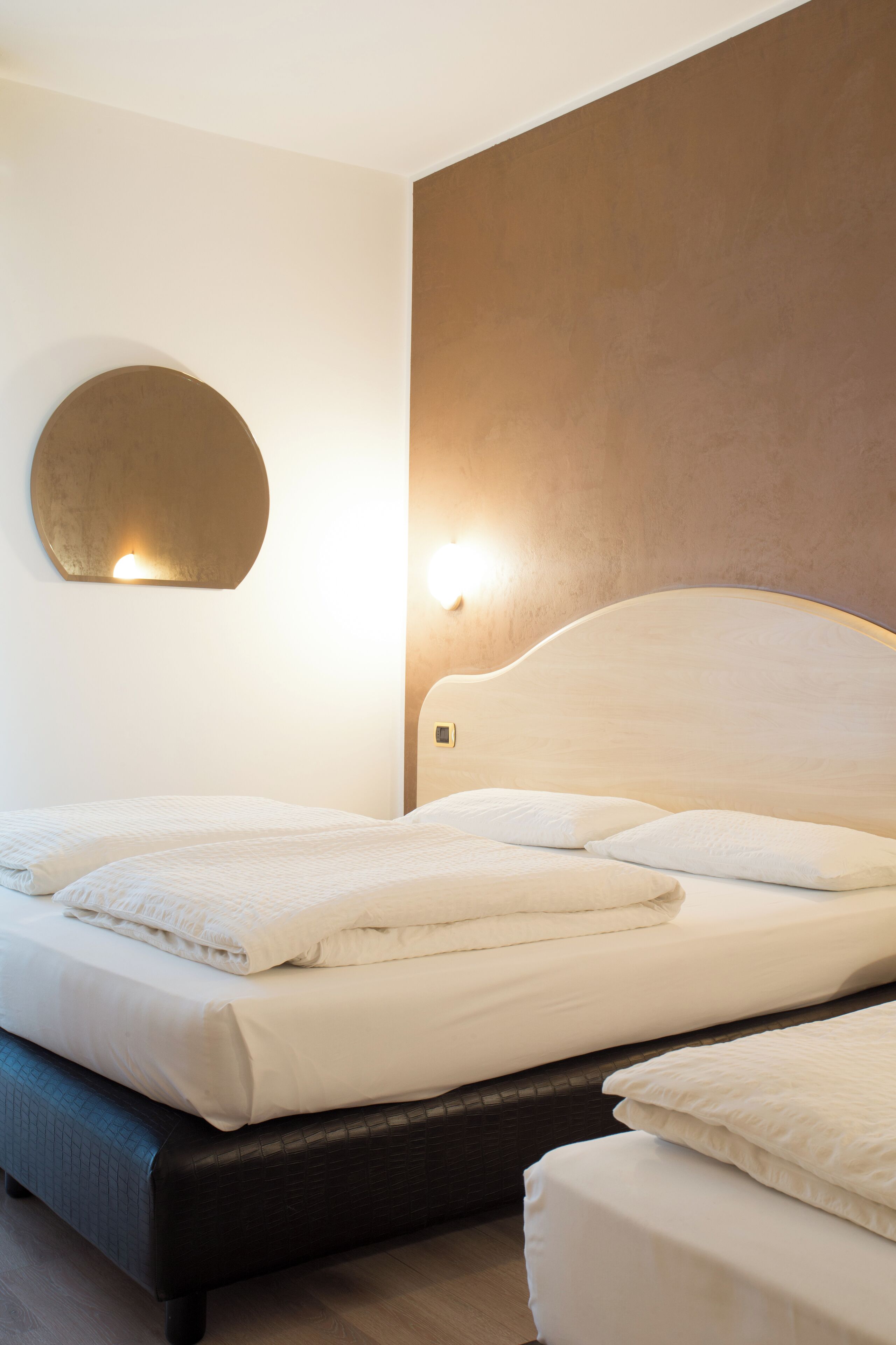Quarto de hotel com uma cama, um espelho redondo e uma luminária fixada na parede.