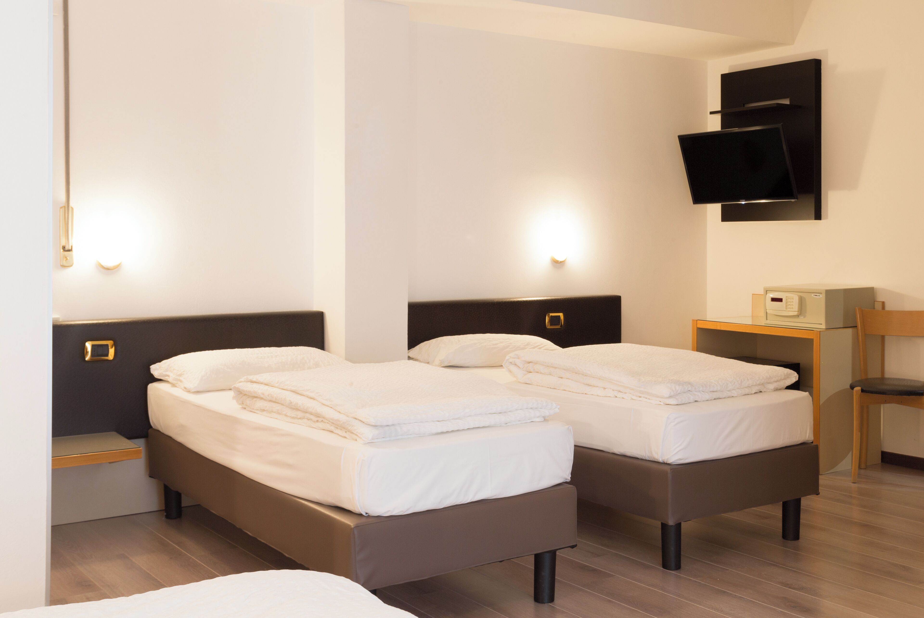 A hotel room with two beds, a TV mounted on the wall, a small desk with a chair, and a bedside table with a lamp.