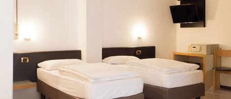 A hotel room with two beds, a TV mounted on the wall, a small desk with a chair, and a bedside table with a lamp.