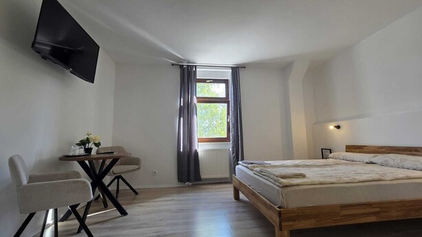 A bedroom with a bed, a desk with a chair, a TV mounted on the wall, and a window with curtains. - Pension Wolf (Hohe Börde)