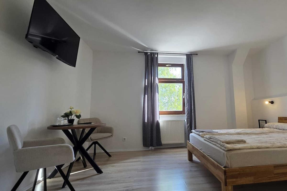 A bedroom with a bed, a desk with a chair, a TV mounted on the wall, and a window with curtains.