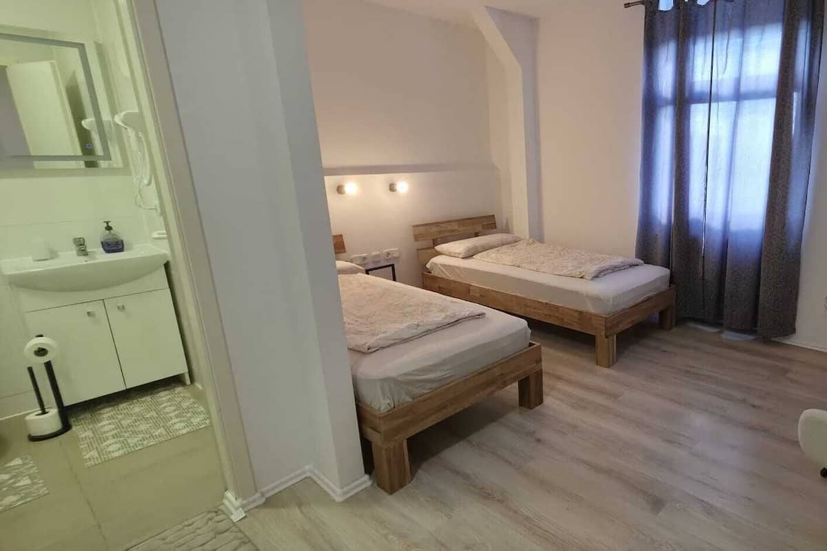A bedroom with two wooden beds, a small table, a chair, a bathroom with a sink and mirror, and a window with curtains.