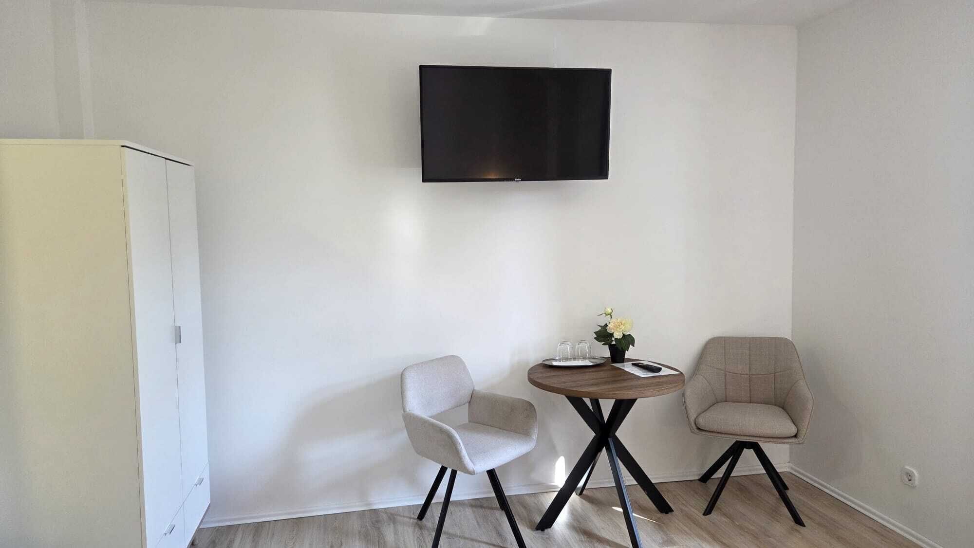 Una habitación con televisión de pantalla plana en la pared, una mesita redonda con dos sillas y un armario.
