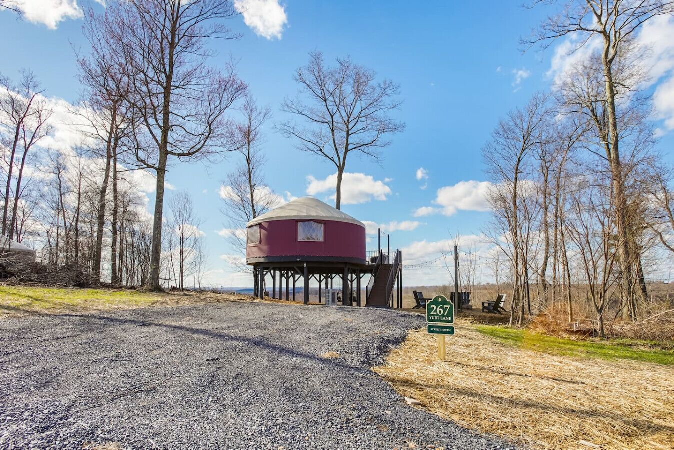 A yurt with a red exterior and a white roof, situated on stilts in a grassy area with trees in the background. A sign indicates the location as '267 Yurt Lane'.