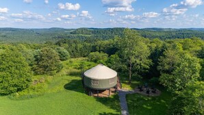 A yurt with a circular roof, surrounded by lush greenery and trees, set in a hilly landscape. - Adventure Abode | Modern Yurt w/ Hot Tub, Bubble Dome + ADA Access (Reynoldsville)