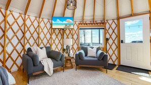 A cozy interior with a wooden lattice wall, two grey sofas, a small round table with a lamp, and a TV screen displaying a beach scene. - Adventure Abode | Modern Yurt w/ Hot Tub, Bubble Dome + ADA Access (Reynoldsville)