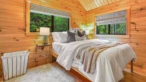 Ein gemütliches Schlafzimmer in einer Holzhütte mit einem Bett, Nachttischen und einem Fenster mit Blick auf Grünflächen.