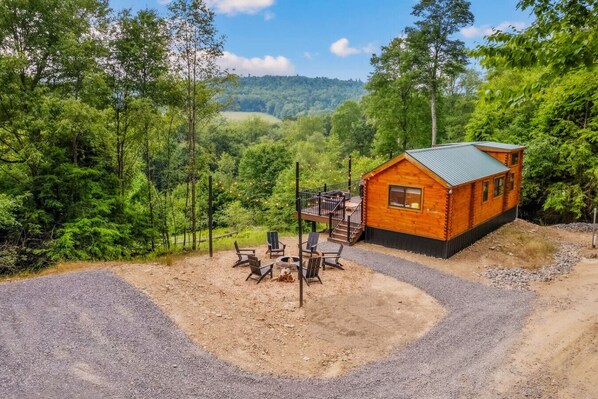 A cabin with a deck, surrounded by a forest and a gravel area with outdoor seating.