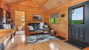 A cozy cabin interior with a wooden floor, a sofa, a dining table, and a kitchen area. - Hidden Creek Hideaway - Cozy Log Cabin w/ Hot Tub, Fire Pit & Scenic Views (Shippenville)