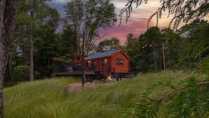 A rustic cabin with a deck, surrounded by tall trees and a grassy field. - Hidden Creek Hideaway - Cozy Log Cabin w/ Hot Tub, Fire Pit & Scenic Views (Shippenville)