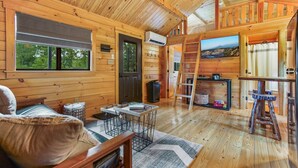 A cozy cabin interior with a wooden interior, a fireplace, a dining area, and a large window overlooking the outdoors. - Hidden Creek Hideaway - Cozy Log Cabin w/ Hot Tub, Fire Pit & Scenic Views (Shippenville)