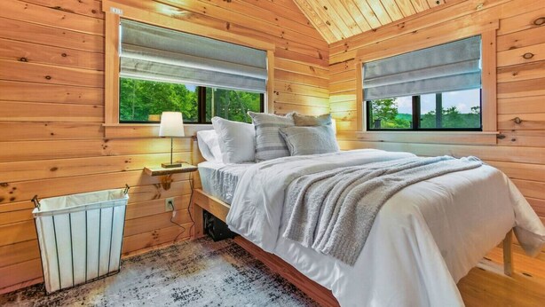 A wooden cabin bedroom with a bed, nightstand, lamp, and a view of greenery through the window. - Hidden Creek Hideaway - Cozy Log Cabin w/ Hot Tub, Fire Pit & Scenic Views (Shippenville)