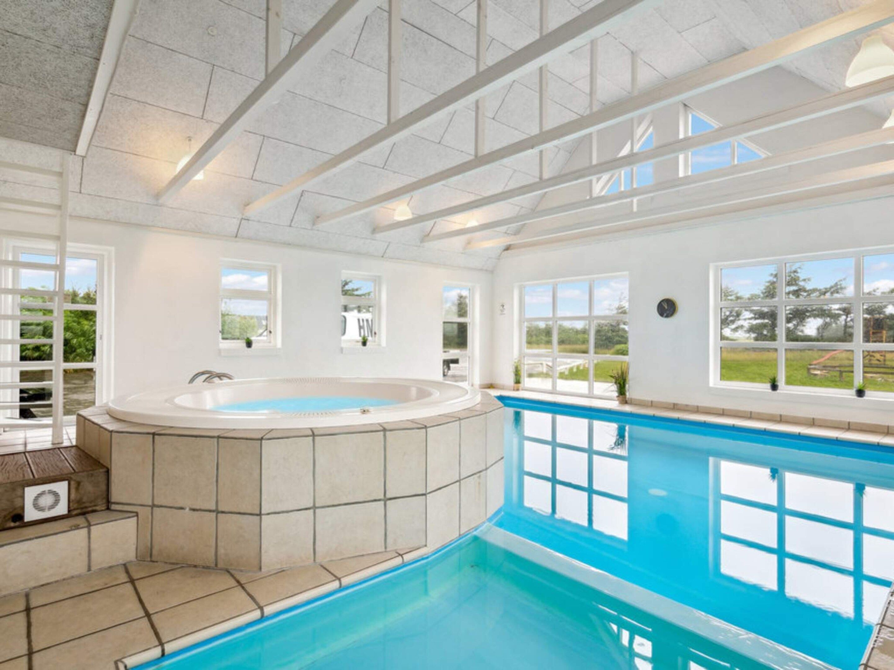 A large indoor swimming pool with a hot tub, surrounded by large windows offering views of the outdoors.