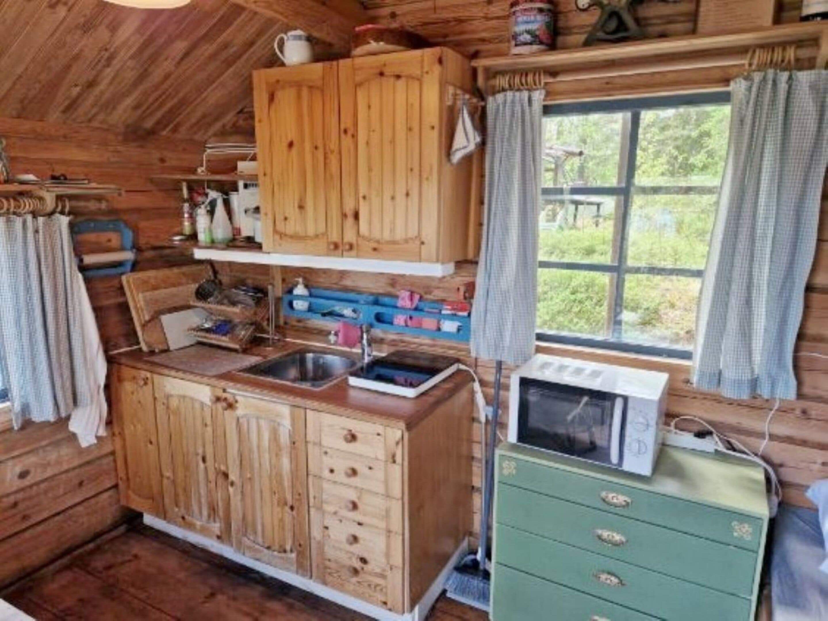 A rustic wooden cabin interior with a kitchenette, wooden cabinets, a green dresser, and a window with curtains.