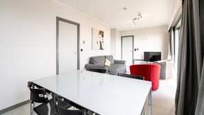 Een moderne woonkamer met een witte eettafel, zwarte stoelen, een grijze bank en een rode fauteuil. Er is een flatscreen televisie op een standaard, een schilderij aan de muur en een raam met gordijnen.