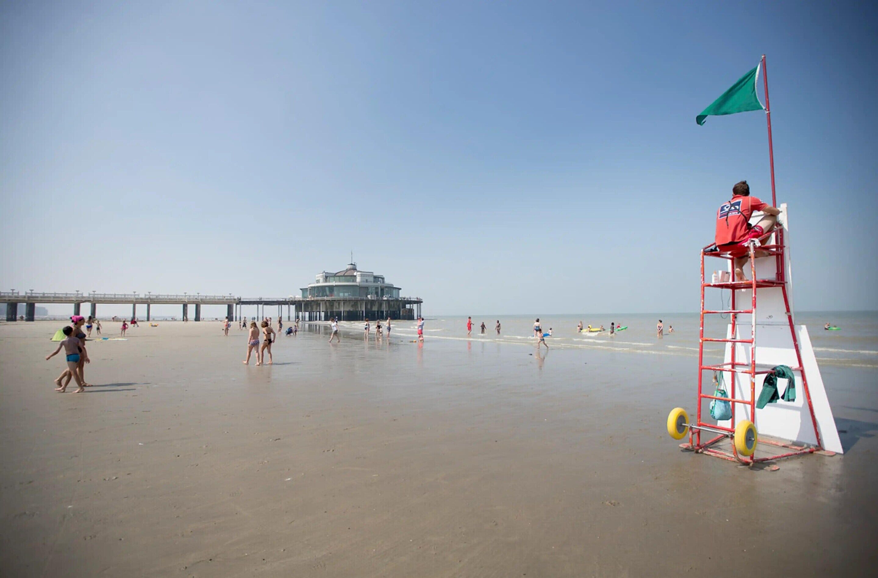 Una playa con una torre de salvavidas, un muelle al fondo y gente en la arena.