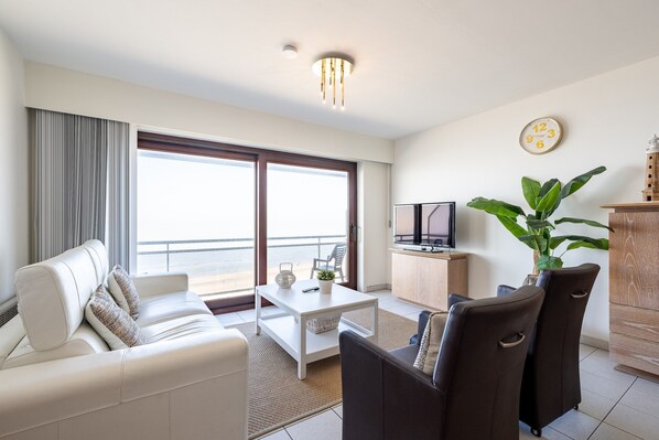 Un salon moderne avec un canapé blanc, une table basse et un téléviseur. Une grande fenêtre avec des rideaux, une plante et une horloge ornent le mur.