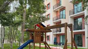 A playground with a slide and swings, surrounded by trees and a building with balconies. - Modern apart in Cancun, well located, amenities. 2 bedrooms and 2 full bathrooms (Cancún)