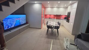 A modern living room with a flat-screen TV, a dining area with a table and chairs, a kitchen with white cabinets and red accents, and a staircase with wooden railings. - Villa with private pool (Orihuela Costa)