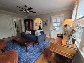 Un salon avec un canapé bleu foncé, une table basse en bois, un tapis bleu à motifs, une table d’appoint en bois avec une lampe et un ventilateur de plafond.