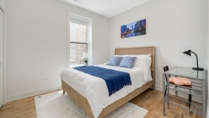 A bedroom with a bed, a window, a desk with a lamp, and a cityscape painting on the wall.