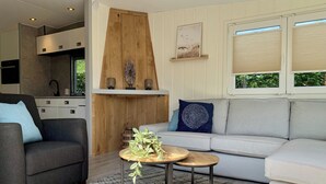 Una sala de estar moderna con un sofá gris, un sillón gris y una pequeña mesa redonda con una planta. Se aprecia una pared de acento de madera con objetos decorativos y una ventana con persianas.