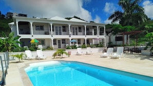 A pool area with lounge chairs and umbrellas, a building with multiple balconies, and lush greenery in the background. - Belvedere Holiday Home (St. John's)