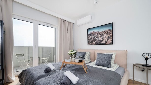 A modern bedroom with a large bed, a balcony with a chair, and a cityscape view.