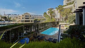 Zona de piscina con sillones de descanso, cielo despejado y edificios modernos al fondo.