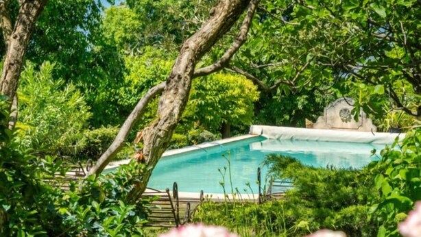 Ein Schwimmbecken, umgeben von üppiger Vegetation und einem Steinbau im Hintergrund.