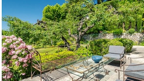 Eine Gartenpatio mit Metallstühlen und einem Glastisch, umgeben von üppigem Grün und blühenden Pflanzen.